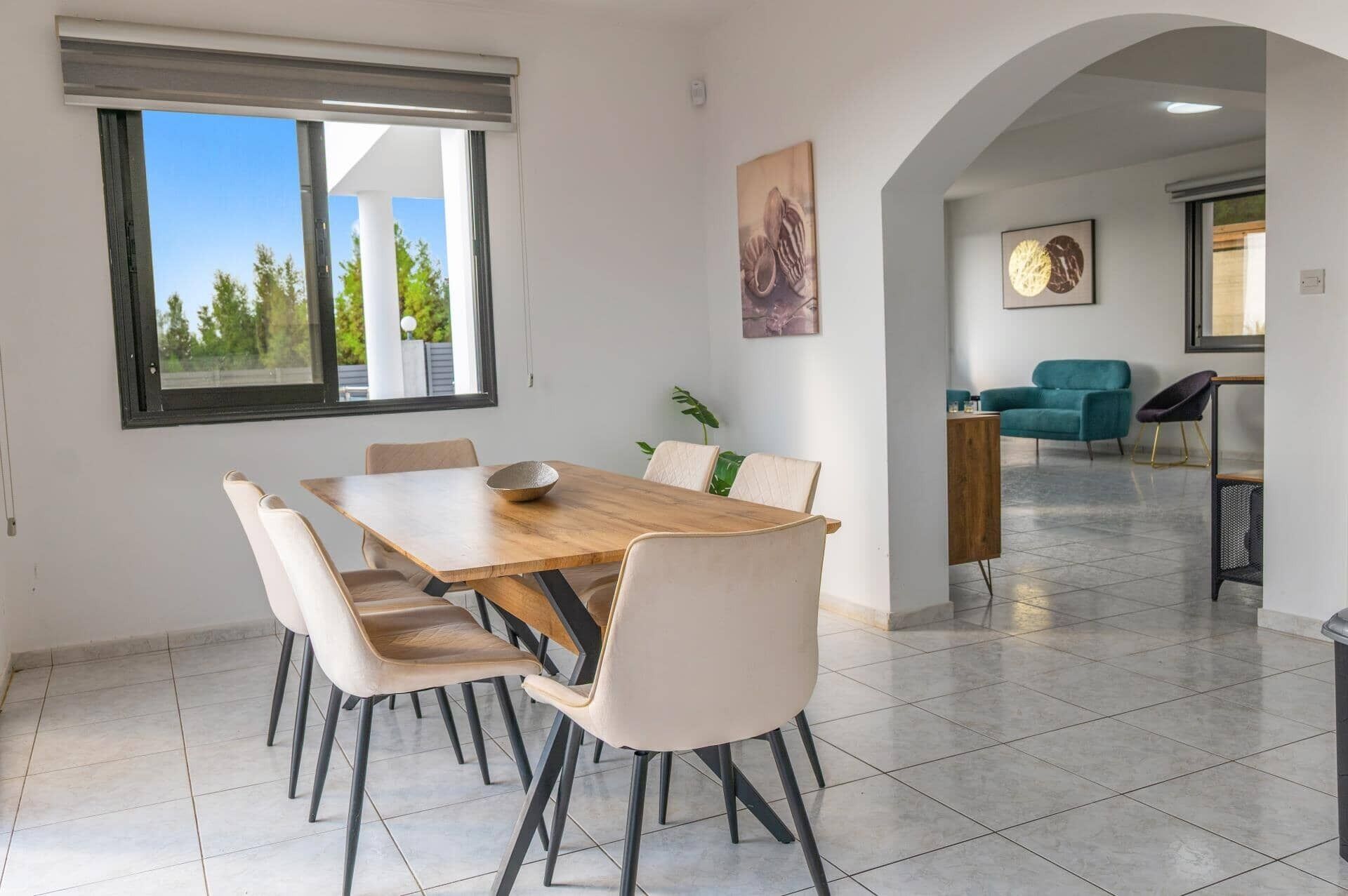 A dining area with a wooden table and six chairs, a large window, and a view of trees.