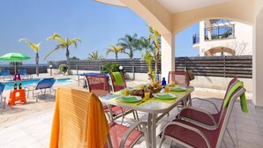 A poolside dining area with a table set for two, chairs, and a view of the pool and palm trees. - Villa Kassiopeia by Villa Plus (Peyia)