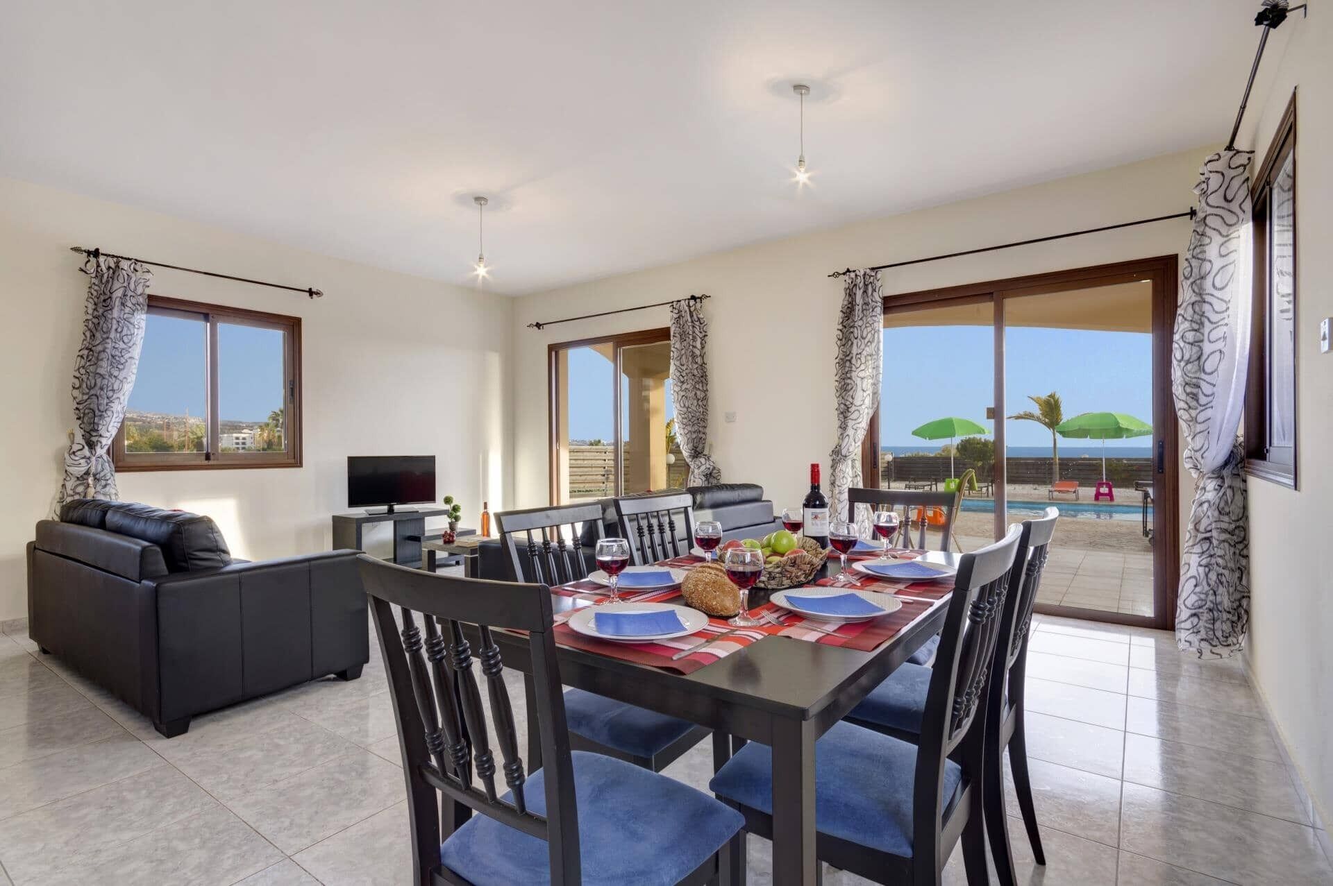 A dining area with a table set for four, a sofa, a TV, and a view of the pool and beach.