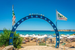 Une entrée de plage agrémentée d’une arche sur laquelle est inscrit « BIENVENUE », de parasols de plage et de drapeaux.