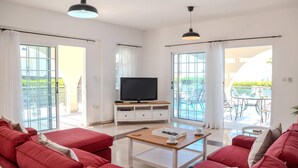 A modern living room with red sofas, a wooden coffee table, a TV on a white cabinet, and large glass doors leading to an outdoor area with a pool.