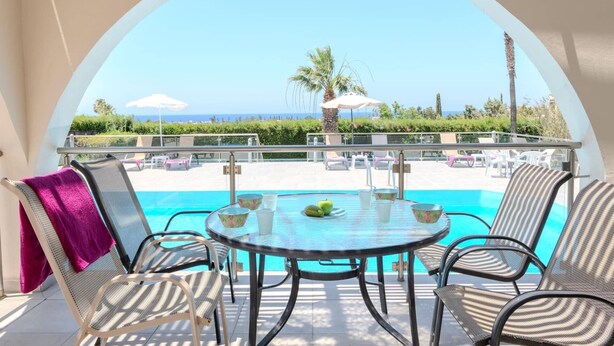 A balcony with a view of a pool and palm trees, a table with chairs, and a purple towel on a chair.
