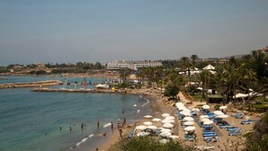 A beach with sun loungers and umbrellas, a large building in the background, and a rocky shoreline.