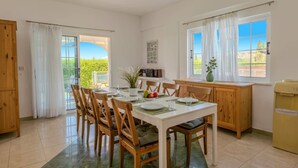 A dining area with a table set for four, wooden chairs, a white cabinet, and a window with a view of greenery. - Villa Joanna Coral Bay by Villa Plus (Peyia)
