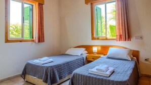 Quarto com duas camas, cada uma com travesseiro e toalha dobrada. Há duas janelas com cortinas e uma mesa de cabeceira com abajur entre as camas.