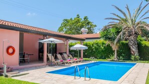 Piscina com espreguiçadeiras e guarda-sóis, cercada por um jardim e um edifício com telhado em azulejos.