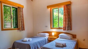 Quarto com duas camas, cada uma com cabeceira de madeira, mesa de cabeceiro e luminária. Janelas com cortinas e piso revestido de azulejos.