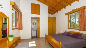 A bedroom with a wooden bed, a bench, a small table, a mirror, and a window with curtains. - Villa Iriss by Villa Plus (Poli Crysochous)