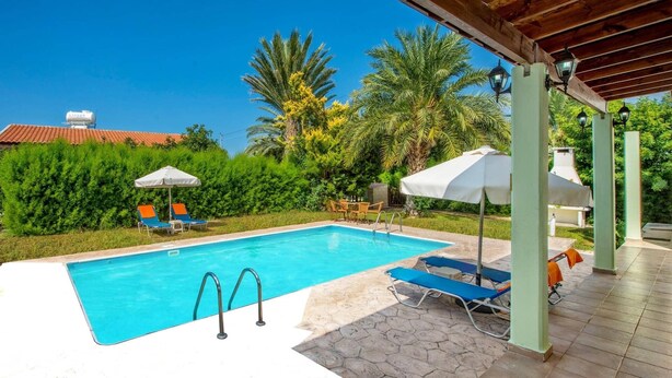 A swimming pool with lounge chairs and umbrellas, surrounded by greenery and a covered area with wooden beams. - Villa Iriss by Villa Plus (Poli Crysochous)