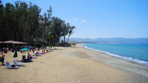 Ein Strand mit Liegestühlen und Sonnenschirmen, einem überdachten Sitzbereich und Blick auf das Meer und sanfte Hügel in der Ferne.