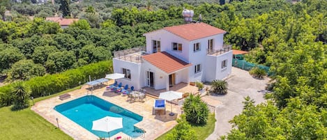 Une maison à deux étages avec une piscine, entourée d’une végétation luxuriante et avec des collines en arrière-plan.