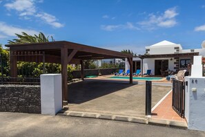Un espacio al aire libre cubierto con piscina, sillas de descanso y un edificio de fachada blanca.