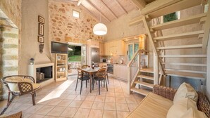 A rustic interior with a stone wall, wooden ceiling, a dining area with chairs, a fireplace, a kitchen with a stove, and a staircase.