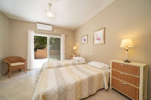 A bedroom with two beds, a wicker chair, a bedside table with a lamp, and a sliding glass door leading to an outdoor area.