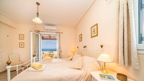 Un dormitorio con dos camas, una mesita de noche con lámpara, una silla y vista al mar a través de una puerta abierta.