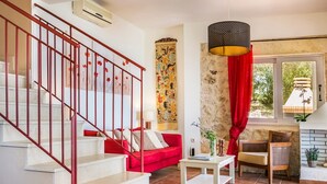 Eine Treppe mit roten Geländern führt zu einem Wohnbereich mit einem roten Sofa, einem weißen Couchtisch und einem roten Sessel. Ein großes Fenster bietet Blick auf viel Grün draußen.