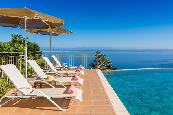 Un espace aménagé au bord de la piscine, avec des chaises longues et un parasol, offrant une vue sur une mer d’un bleu limpide.