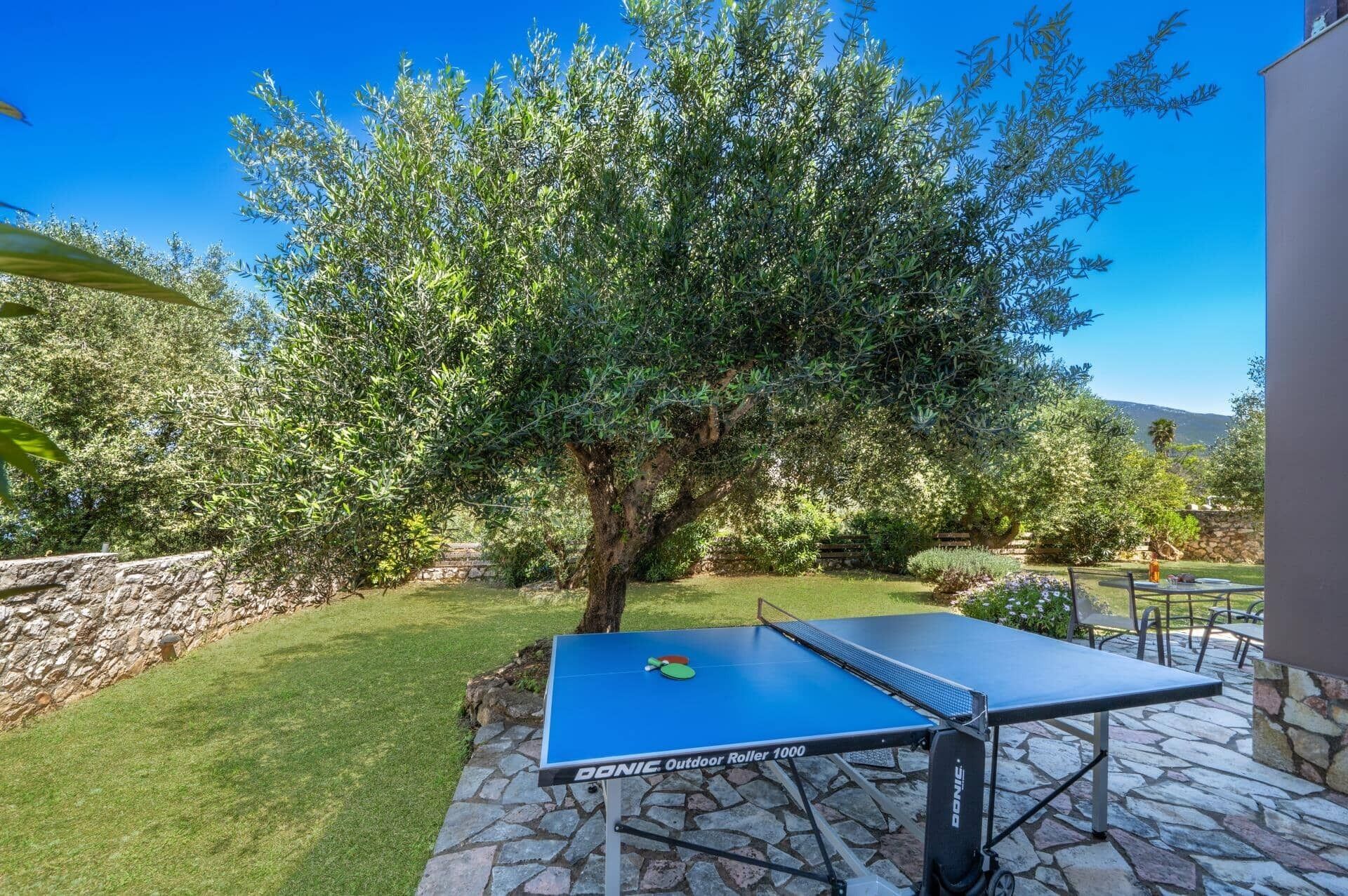 An outdoor ping pong table set up on a stone patio with a large tree in the background.