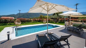 A swimming pool with lounge chairs and umbrellas, surrounded by a landscaped garden and a view of mountains. - Villa Nectar by Villa Plus (Cephalonia)