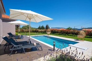 A swimming pool with lounge chairs and umbrellas, surrounded by a landscaped garden and a view of distant hills.