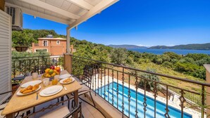 Un balcón con una mesa dispuesta para el desayuno, con vistas a una piscina y un paisaje pintoresco de colinas y una masa de agua.