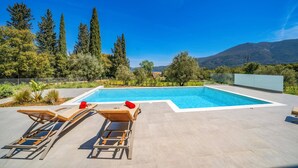 Piscina retangular com duas espreguiçadeiras e divisória de vidro, cercada por árvores e vista para a montanha.
