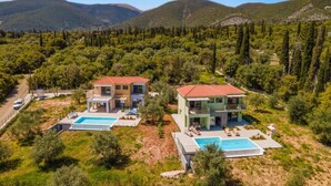 Duas casas modernas com telhados de telha vermelha, cada uma com piscina e rodeadas por vegetação exuberante e montanhas.