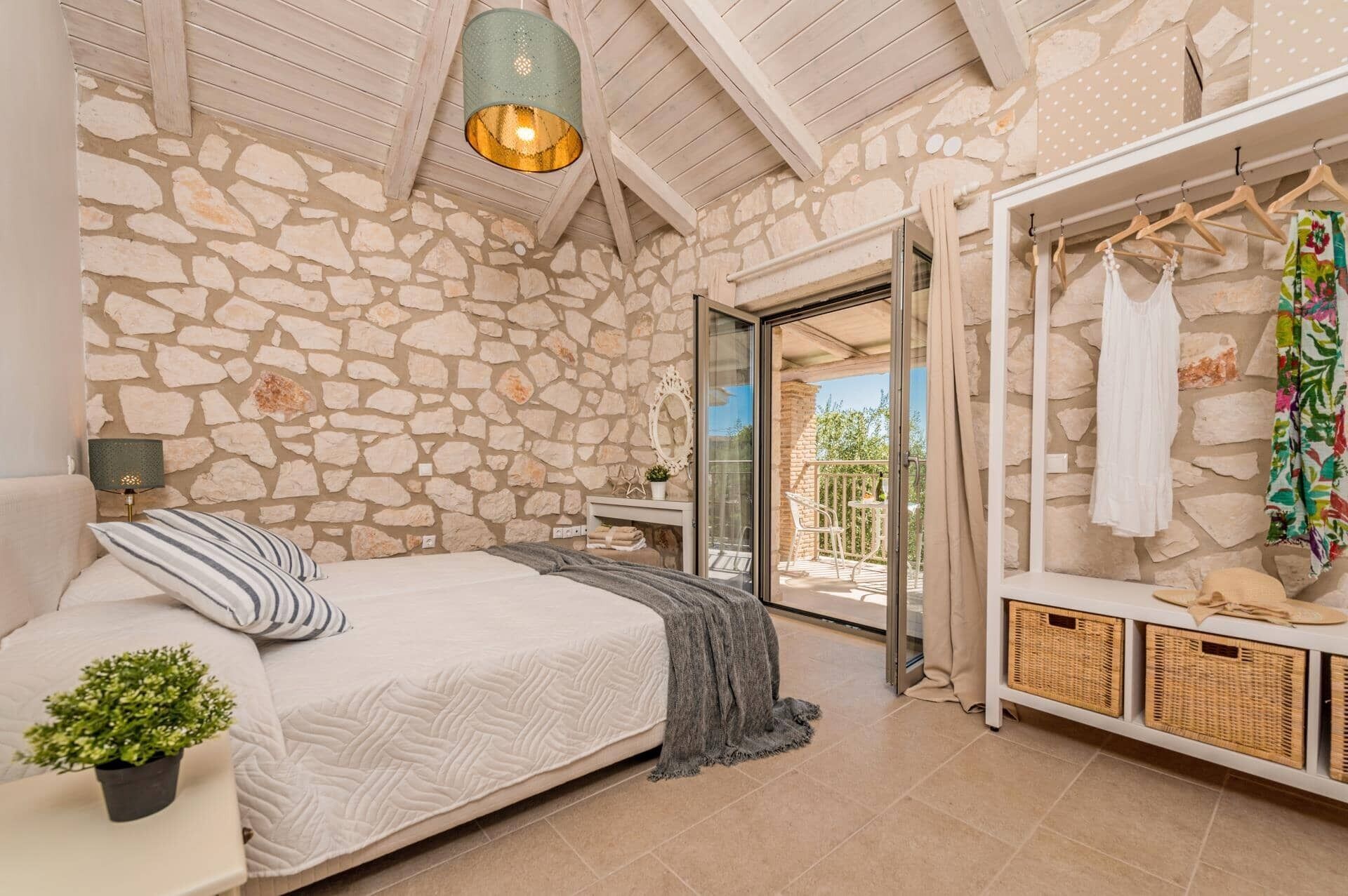 A bedroom with a stone wall, a bed with a striped pillow, a hanging light fixture, and a clothing rack with a white dress.