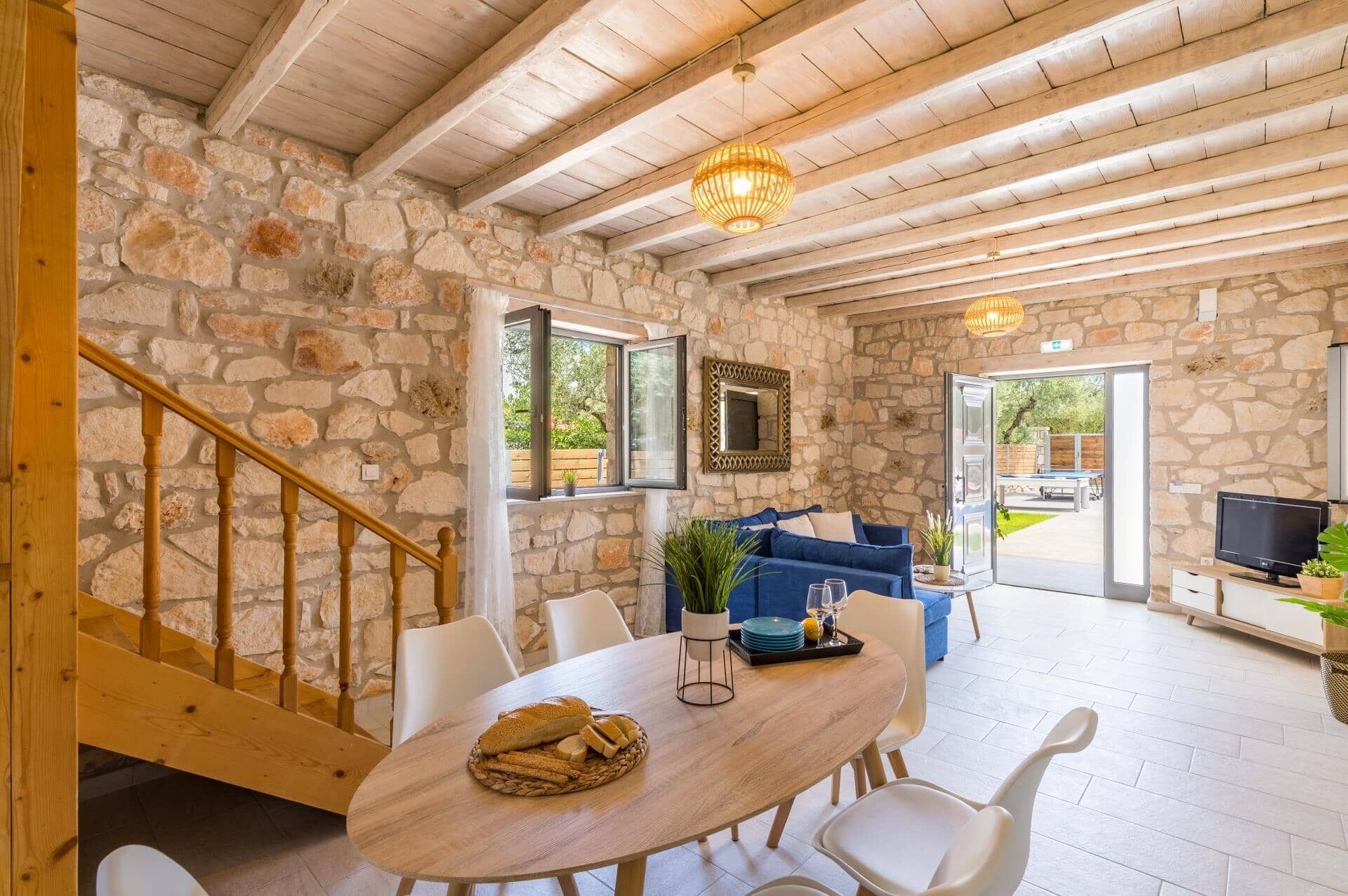 A stone-walled room with a wooden staircase, a dining table with bread, a blue sofa, and a TV.