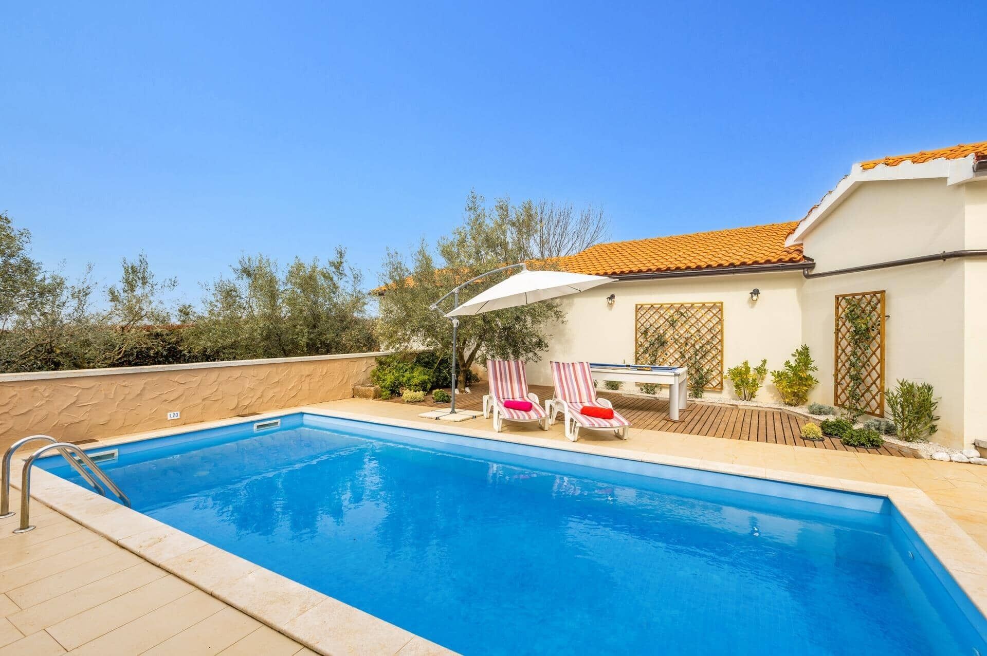 Piscina com espreguiçadeiras e mesa, cercada por um edifício branco com telhado telhado.