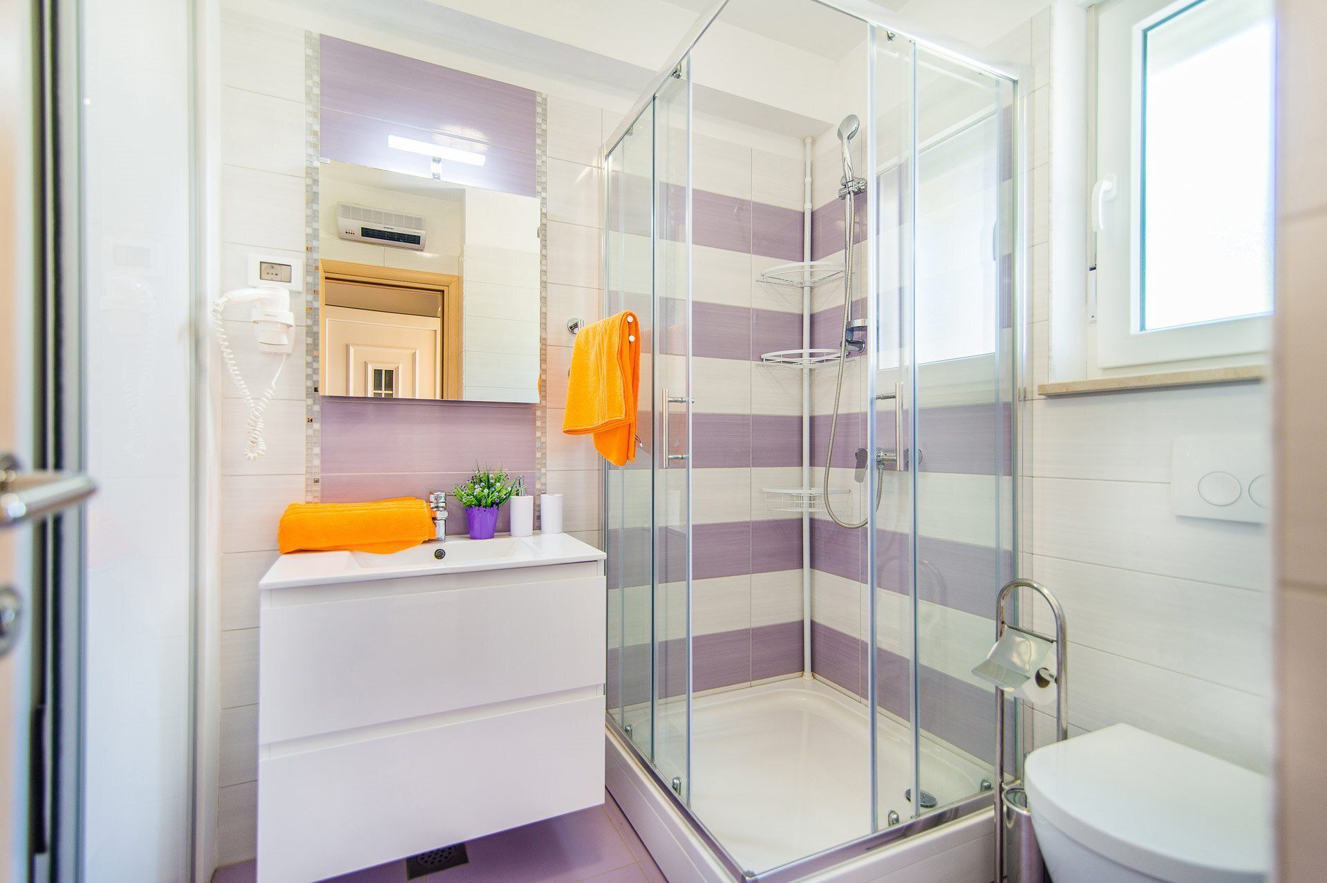 Un baño moderno con cabina de ducha de vidrio, un lavamanos blanco y una planta morada, además de un inodoro.