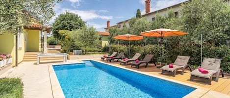 A swimming pool with lounge chairs and umbrellas, surrounded by greenery and a building with a tiled roof.