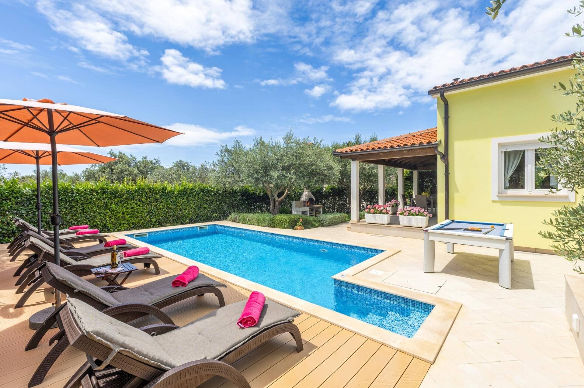Ein Swimmingpool mit Liegestühlen und Sonnenschirm, ein Barbereich und ein Gebäude mit einem Ziegeldach.