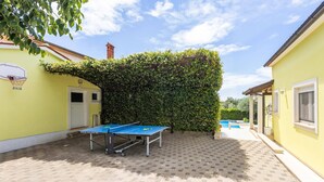 An outdoor table tennis table with a net, set up in a courtyard with a hedge, a basketball hoop, and a pool in the background. - Villa Ladonja by Villa Plus (Pula)