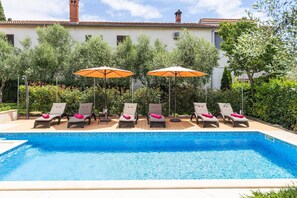 A swimming pool with lounge chairs and umbrellas, surrounded by greenery and a building with a tiled roof. - Villa Ladonja by Villa Plus (Pula)