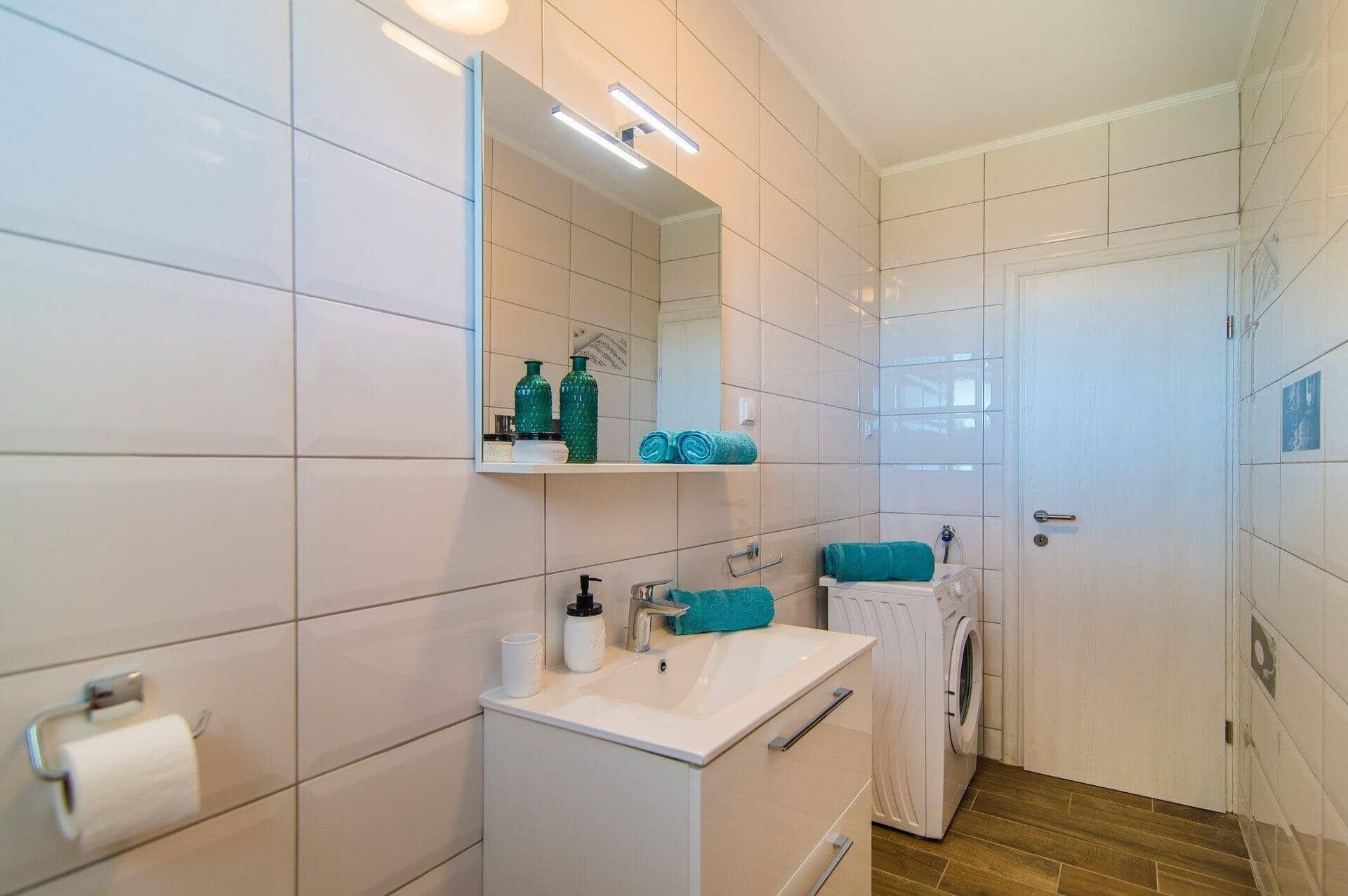 Uma casa de banho moderna com azulejos brancos, um espelho, uma pia, um porta-toalhas e uma máquina de lavar roupa.