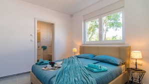Un dormitorio con cama, ventana, mesita de noche y baño visible a través de una puerta abierta.