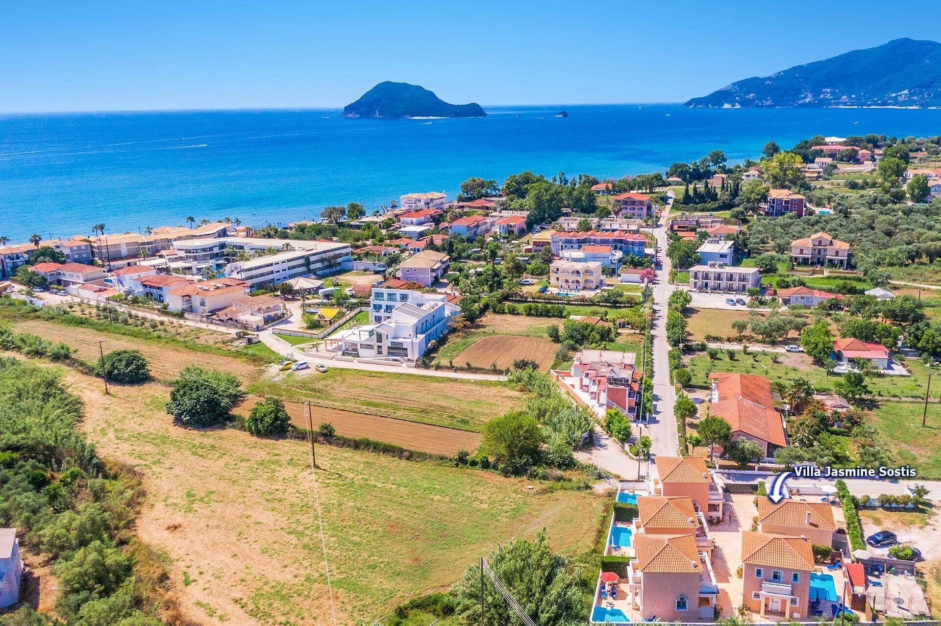 Vue aérienne de la villa Jasmine Sostis, qui offre un panorama sur la côte, avec un mélange de bâtiments résidentiels et d’espaces verts ouverts.