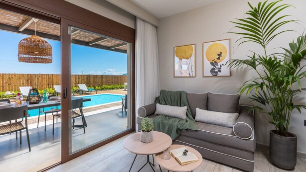 Una sala moderna con un sofá gris, una mesa de centro y una planta grande en maceta. Hay una puerta de cristal que da a una zona de piscina con mobiliario exterior.