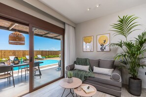 A modern living room with a grey sofa, a coffee table, and a large potted plant. There is a glass door leading to a pool area with outdoor furniture. - Villa Maira Rose by Villa Plus (Katastari)
