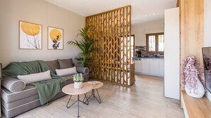 Una sala de estar moderna con un sofá, una mesa de centro y una planta decorativa. Un separador de madera divide la sala de estar del área de la cocina.