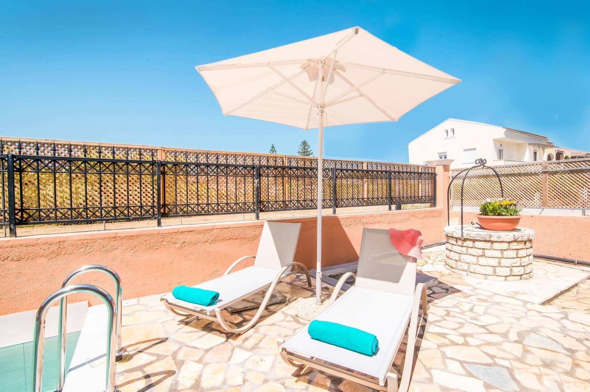 Eine Dachterrasse mit zwei Liegestühlen, einem großen Sonnenschirm, einem kleinen Brunnen und einem Poolbereich.