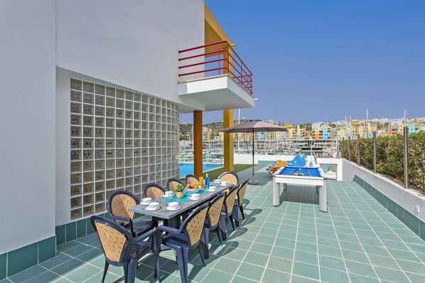 A rooftop terrace with a pool table, dining table, and chairs, overlooking a marina with boats and colorful buildings.