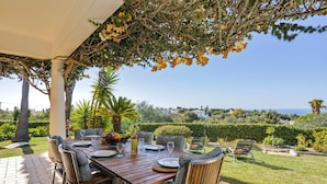 An outdoor dining area with a wooden table and chairs, surrounded by greenery and a view of the sea. - Villa Maisie by Villa Plus (Praia do Carvoeiro)