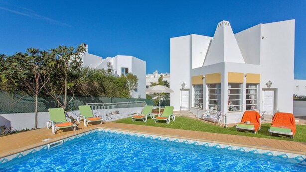 Una piscina con sillas de descanso, una zona de estar cubierta y un edificio con fachada blanca.