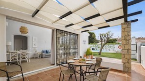 Un patio al aire libre con una mesa redonda y sillas, un espacio de descanso cubierto y vistas a una piscina y follaje.