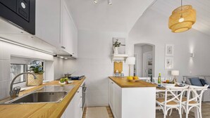 Una cocina moderna con armarios blancos, una encimera de madera y una isla central. Hay una zona de comedor con sillas blancas y una mesa, con una lámpara colgante encima.