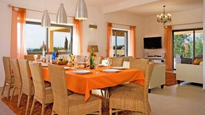 A dining area with wicker chairs and an orange tablecloth, a chandelier, and a television in the living area. - Villa Miranda by Villa Plus (Albufeira)