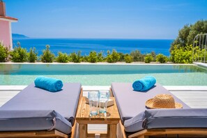 Zona relax a bordo piscina con due lettini, un tavolo con bicchieri e un cappello di paglia, e vista sul mare.