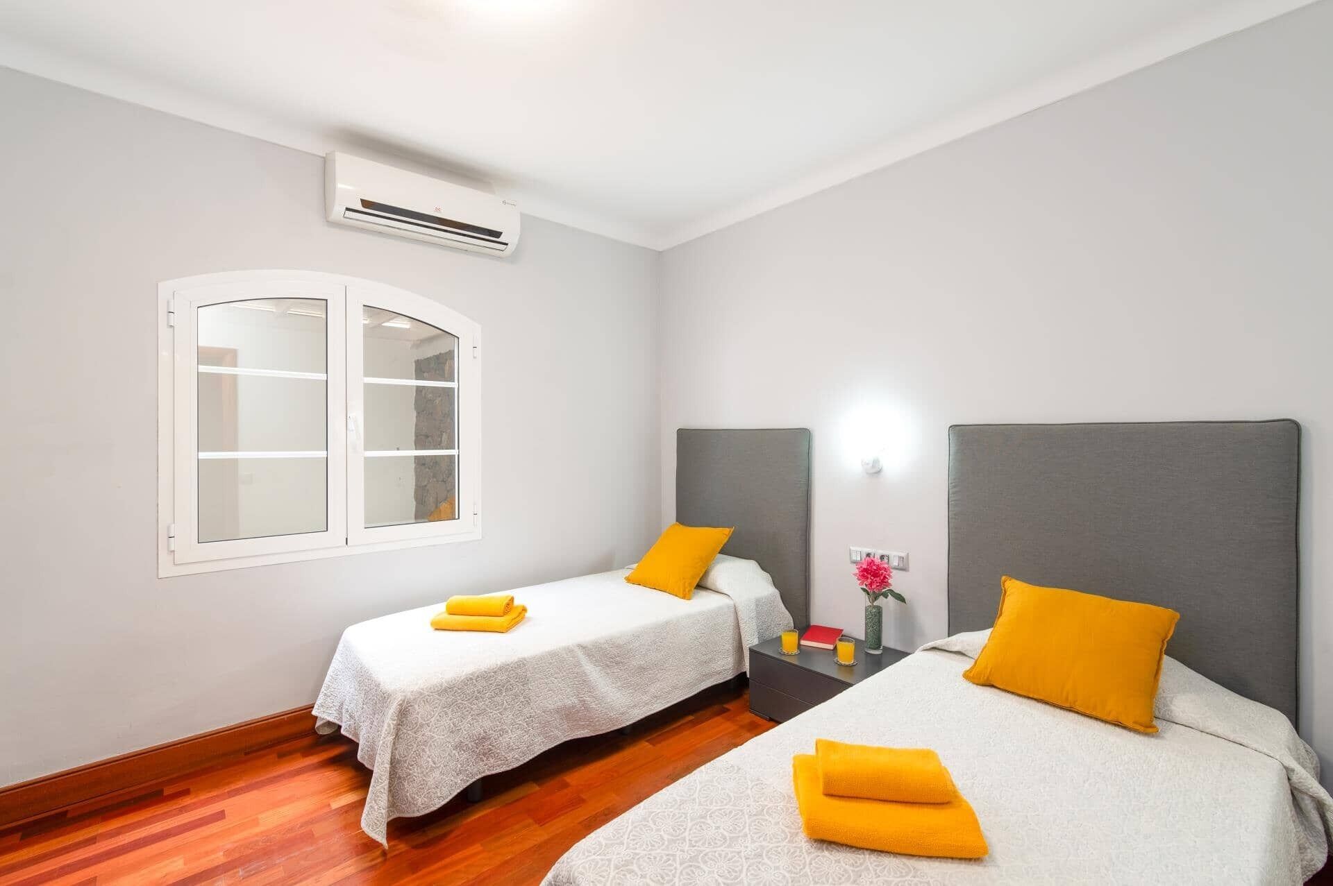 Une chambre d’hôtel avec deux lits, une fenêtre, un climatiseur et une table de chevet sur laquelle se trouvent un livre et un vase de fleurs.